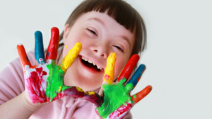 Fotografia przedstawia uśmiechniętą dziewczynkę z Zespołem Downa z pomalowanymi dłońmi farbami.